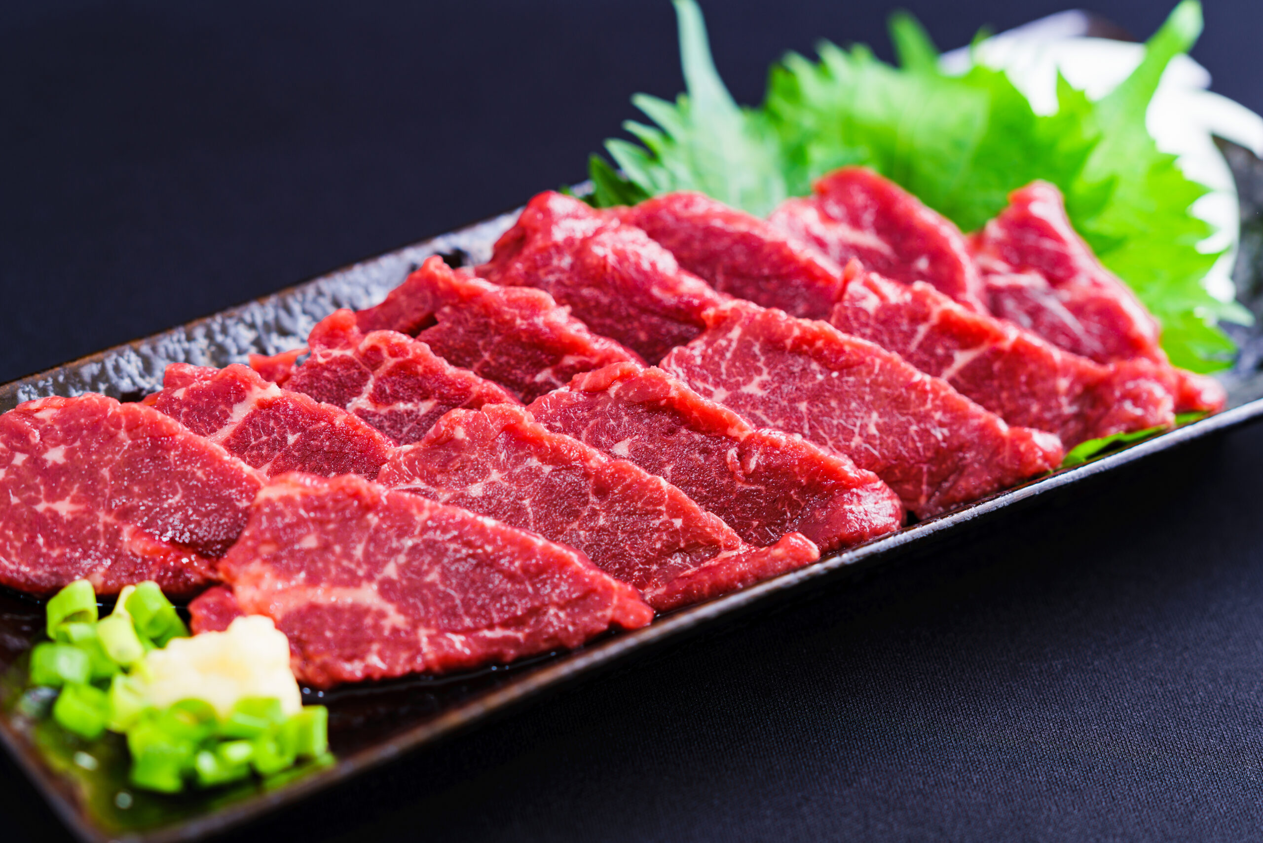 Thinly sliced marbled beef arranged on a black platter with bright green garnish at the edge.