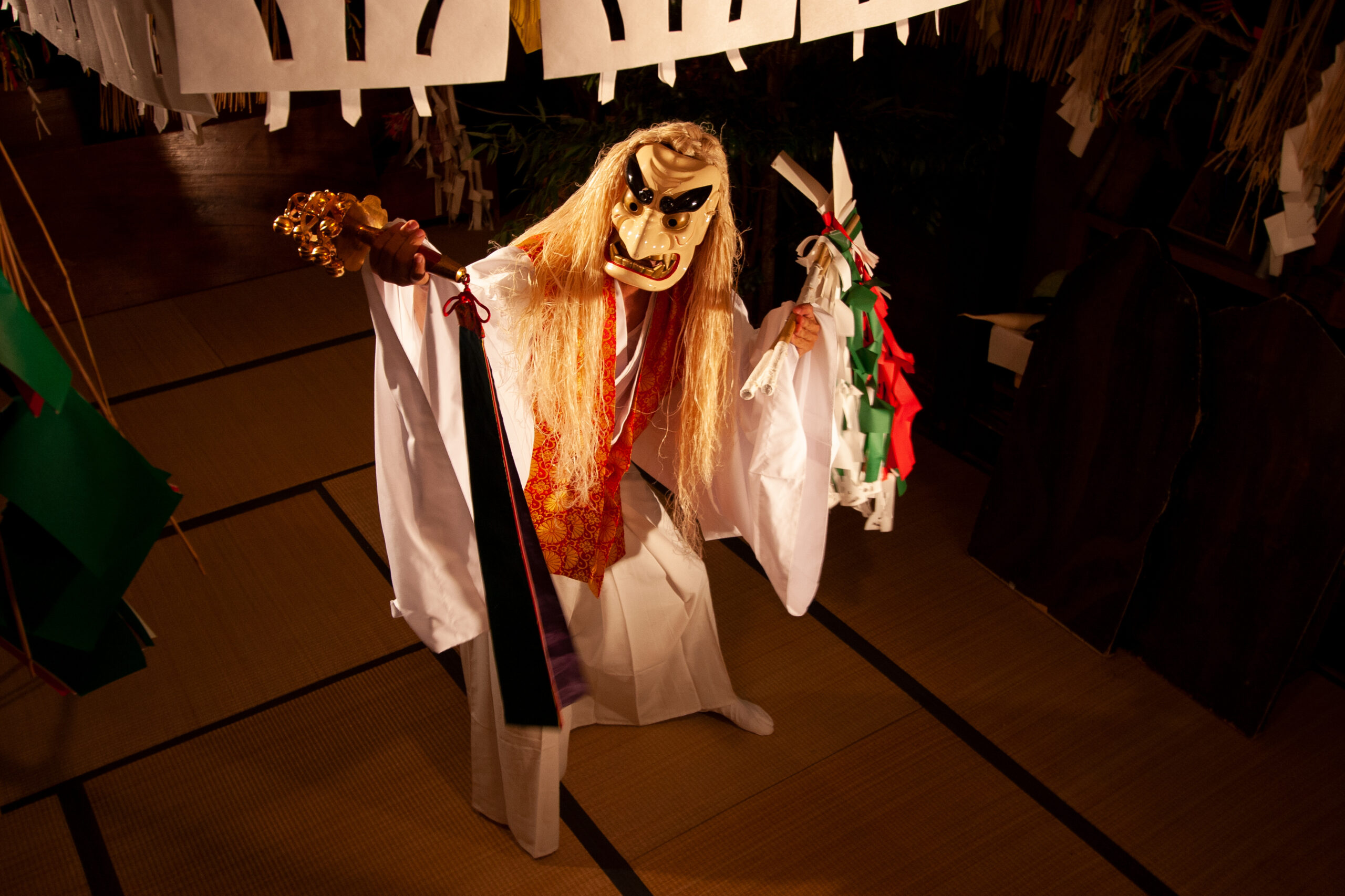 Person in a traditional mask and white robes performing with a golden staff and paper streamers during a ritual scene.