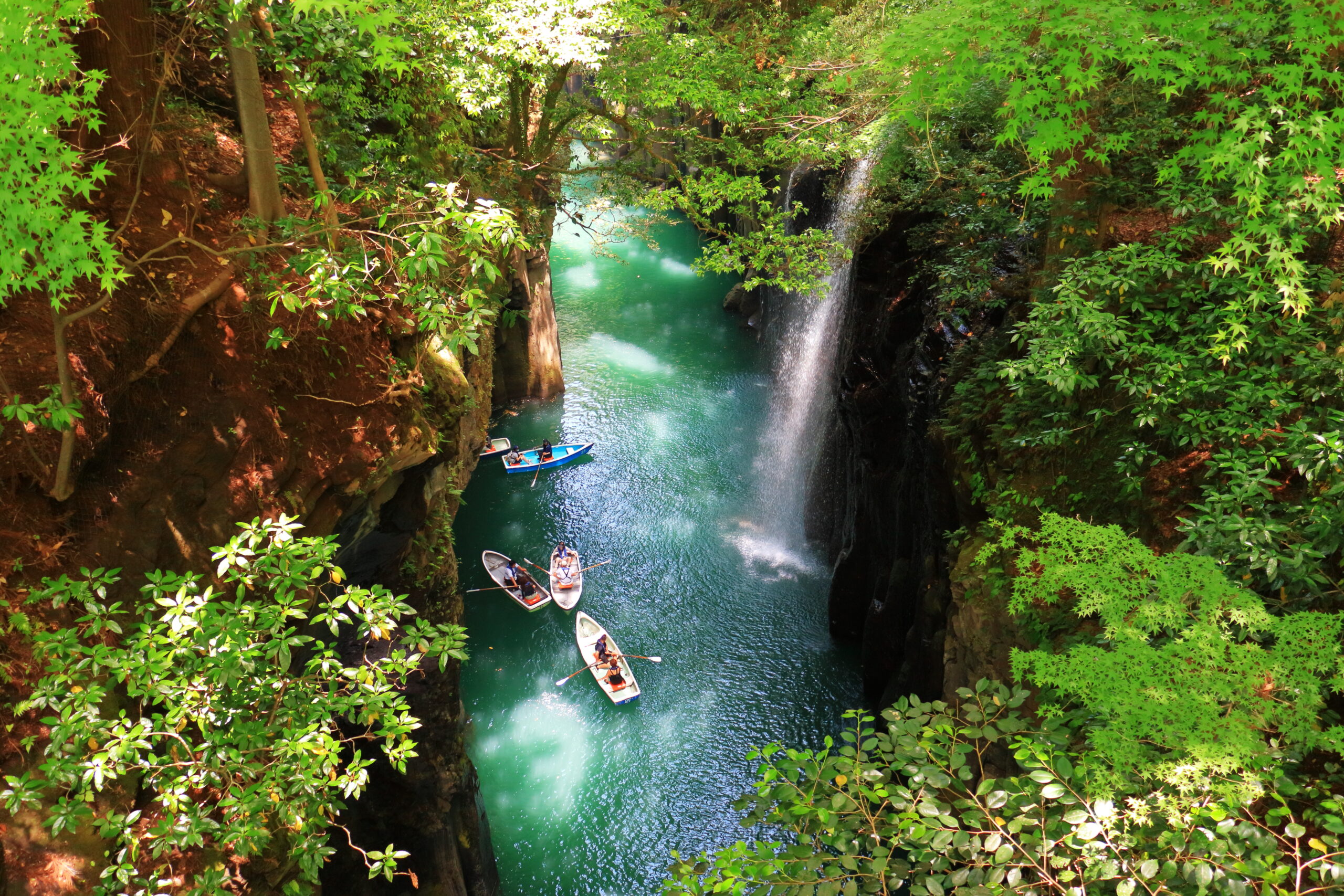 Turquoise river in a canyon, with a waterfall on the right and several small boats paddling among lush green trees overhead.