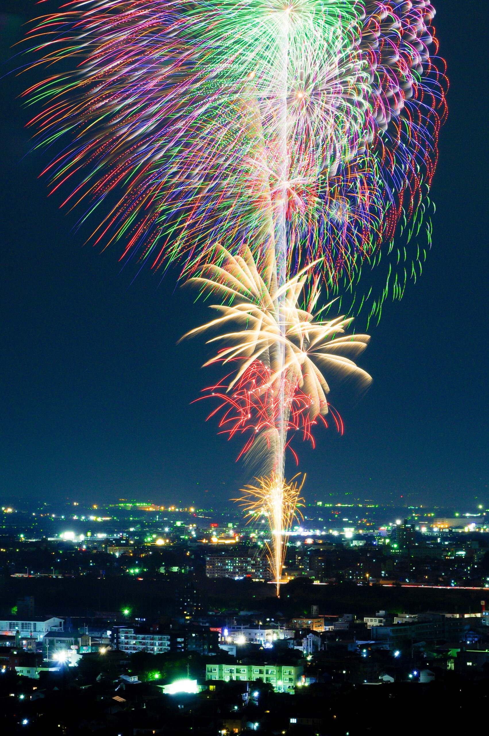 Colorful fireworks exploding in the night sky above a city, with bright green, red, blue, and white trails lighting the scene.