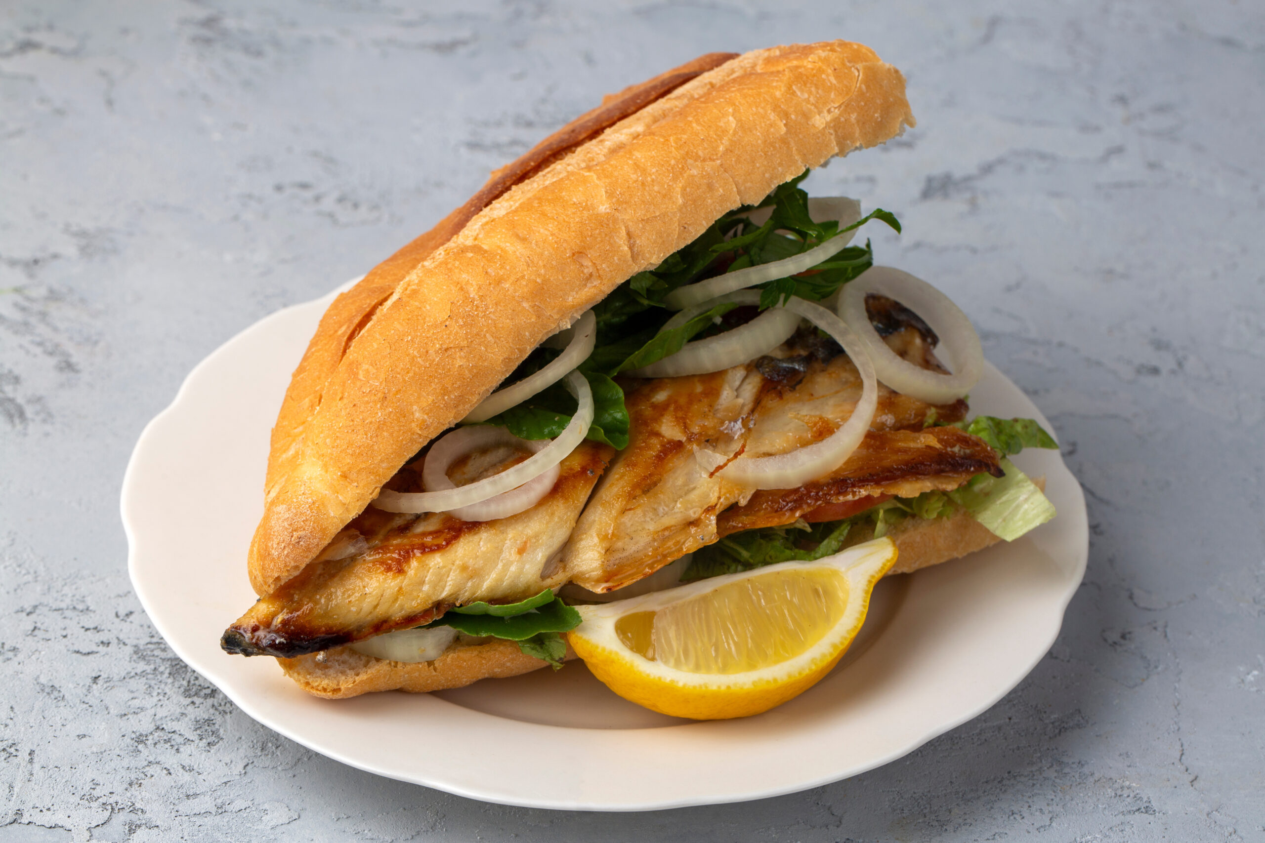 Baguette-style chicken sandwich with lettuce and onion slices on a white plate, lemon wedge nearby.