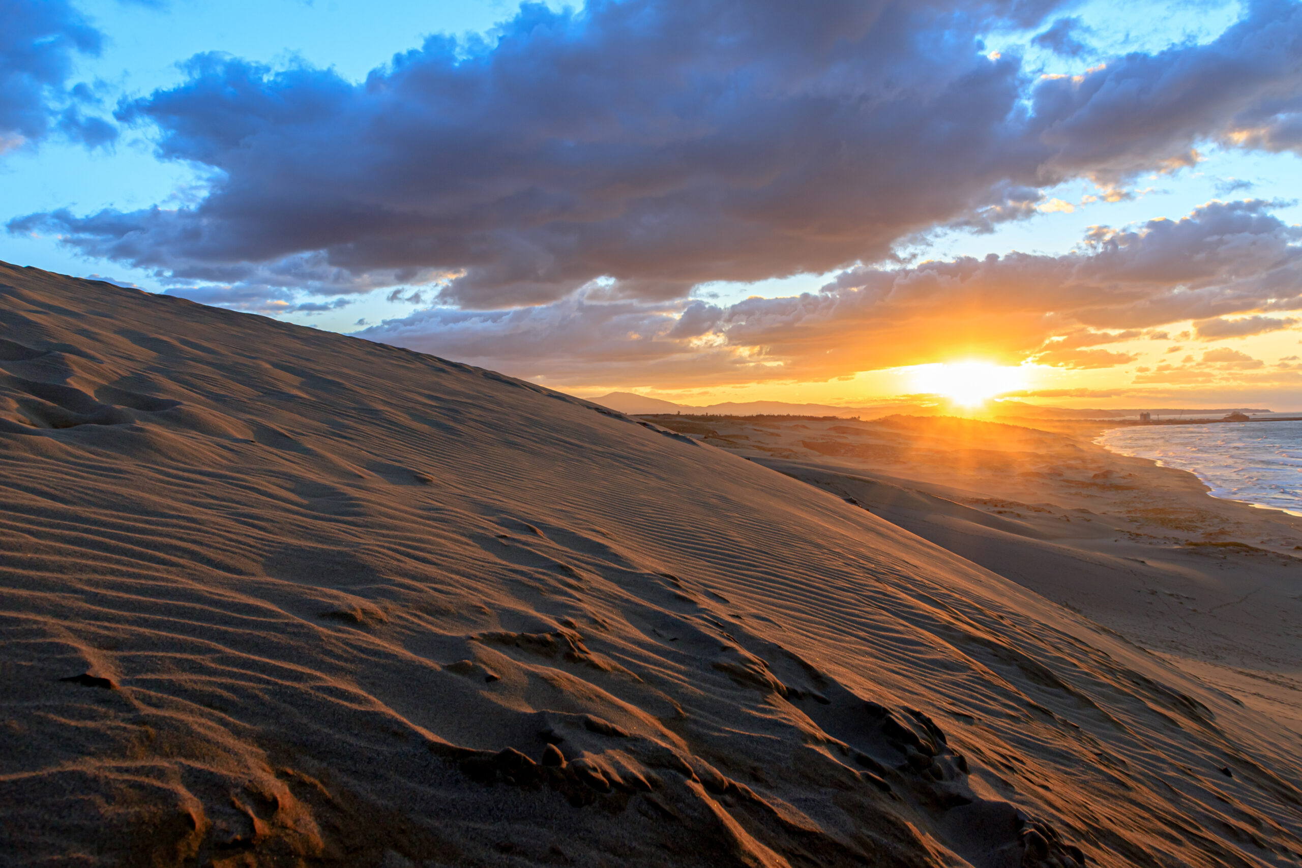 Sunset over rolling sand dunes by the coast, with warm orange light and a blue, cloud-filled sky.