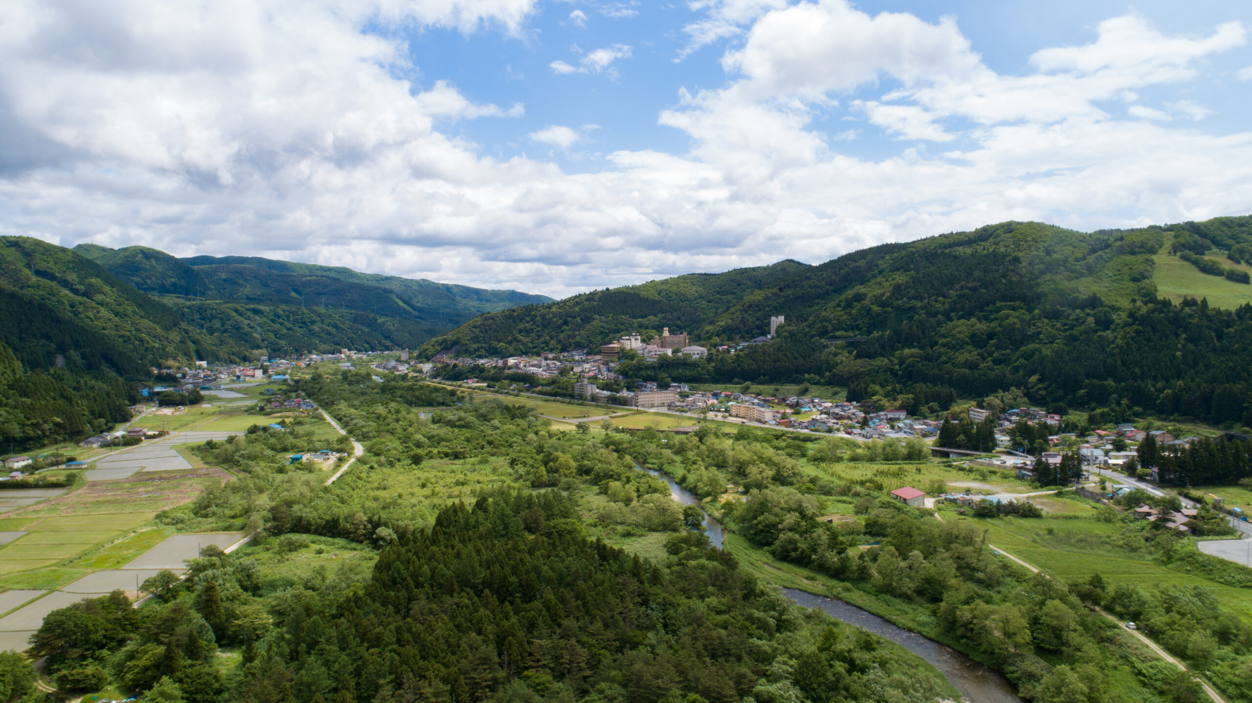 A lush valley town nestled among green hills with a river winding through fields under a cloudy sky.