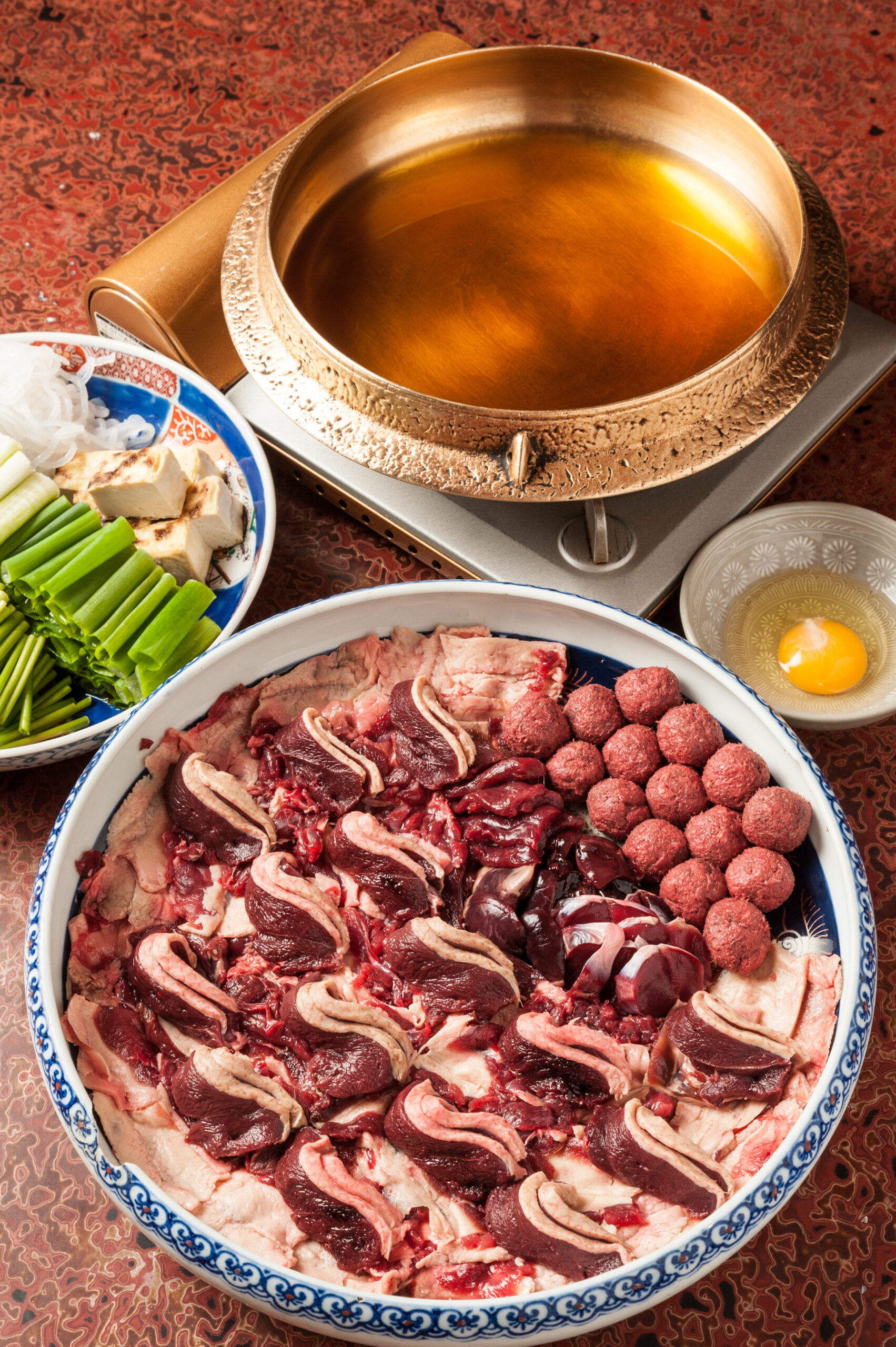 Large blue-and-white bowl filled with thinly sliced beef and other meats arranged for hot pot, with a copper pot nearby.
