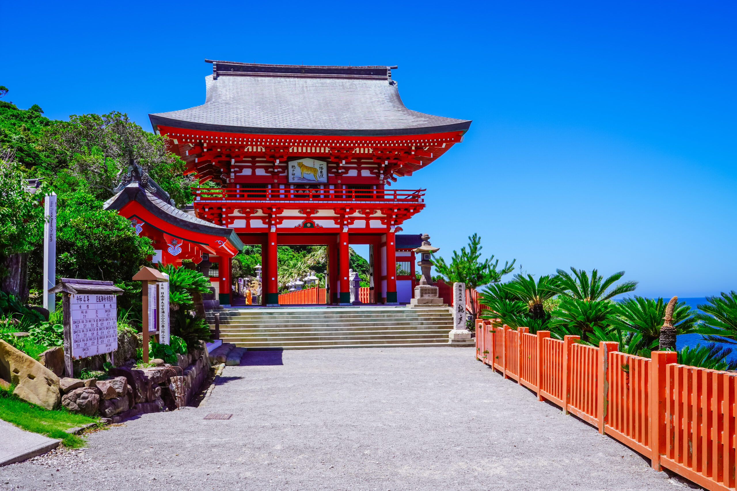 Red temple gate with curved roof and intricate red beams, set among green plants and a bright blue sky.