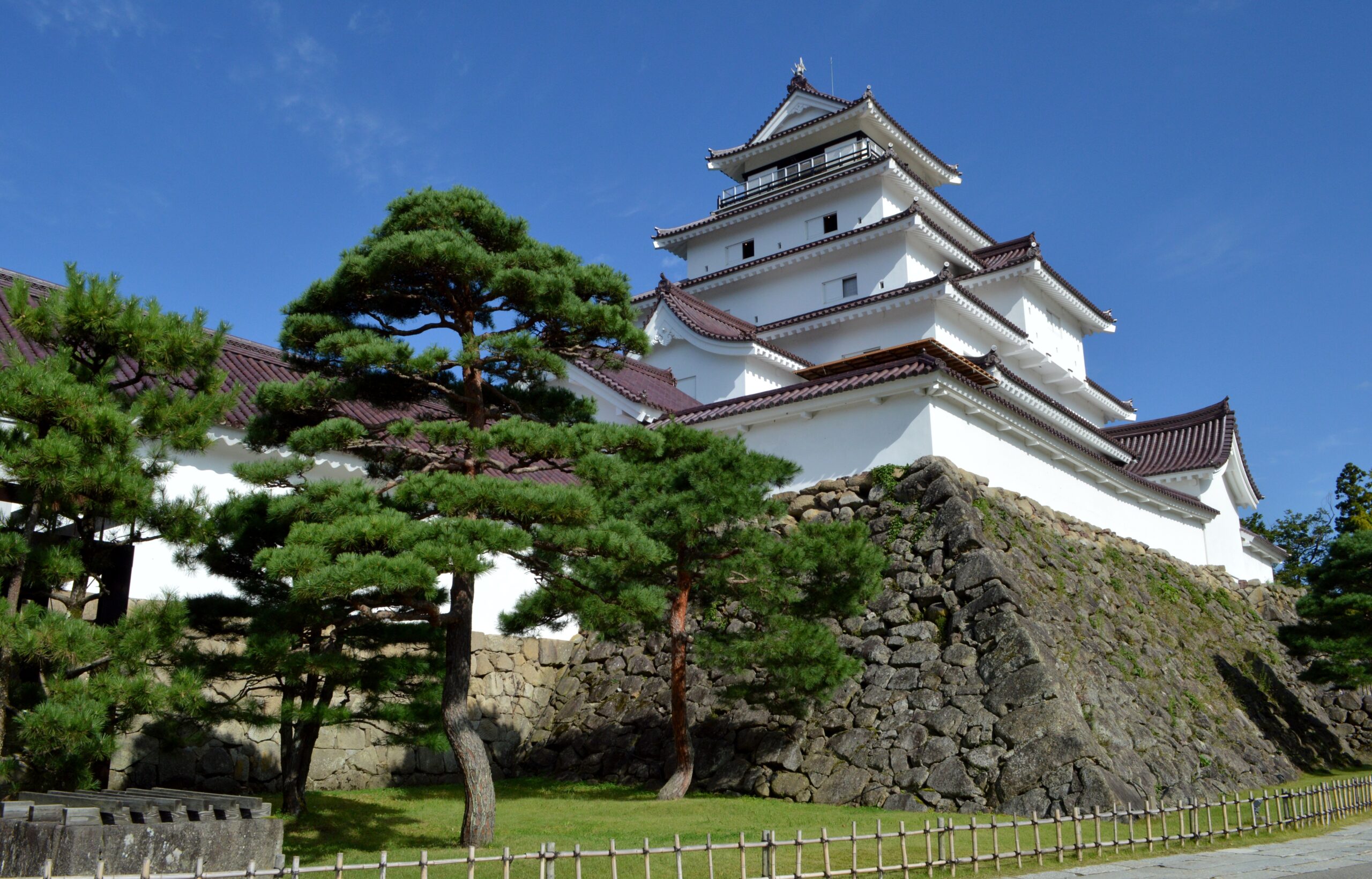 White multi-tier Japanese castle atop a stone retaining wall, with green pines against a blue sky.