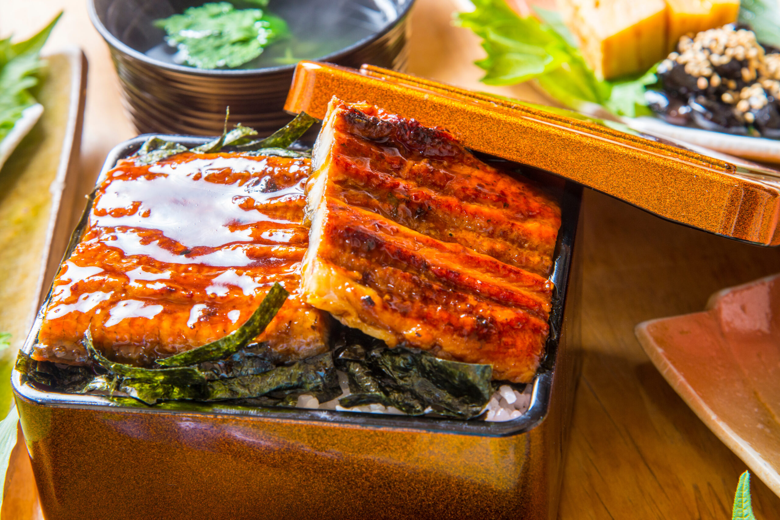 Open lacquered bento box with glossy glazed eel over rice and seaweed, lid resting nearby on a wooden table
