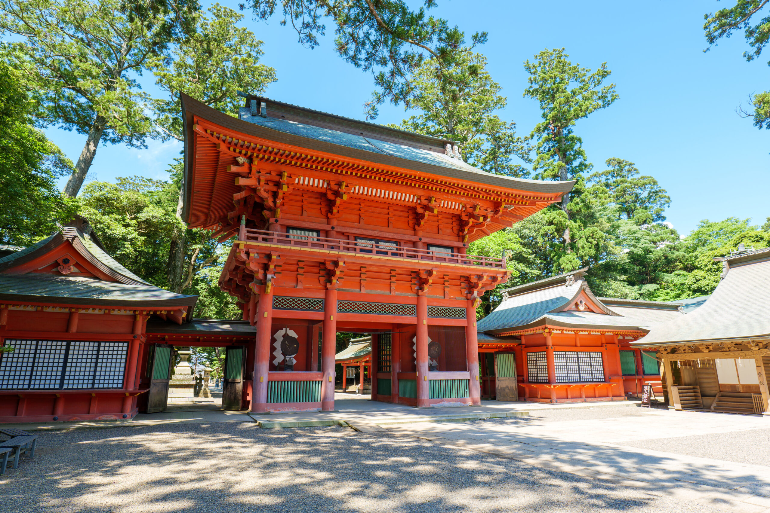Red traditional temple buildings with curved roofs set among tall green trees under a blue sky.
