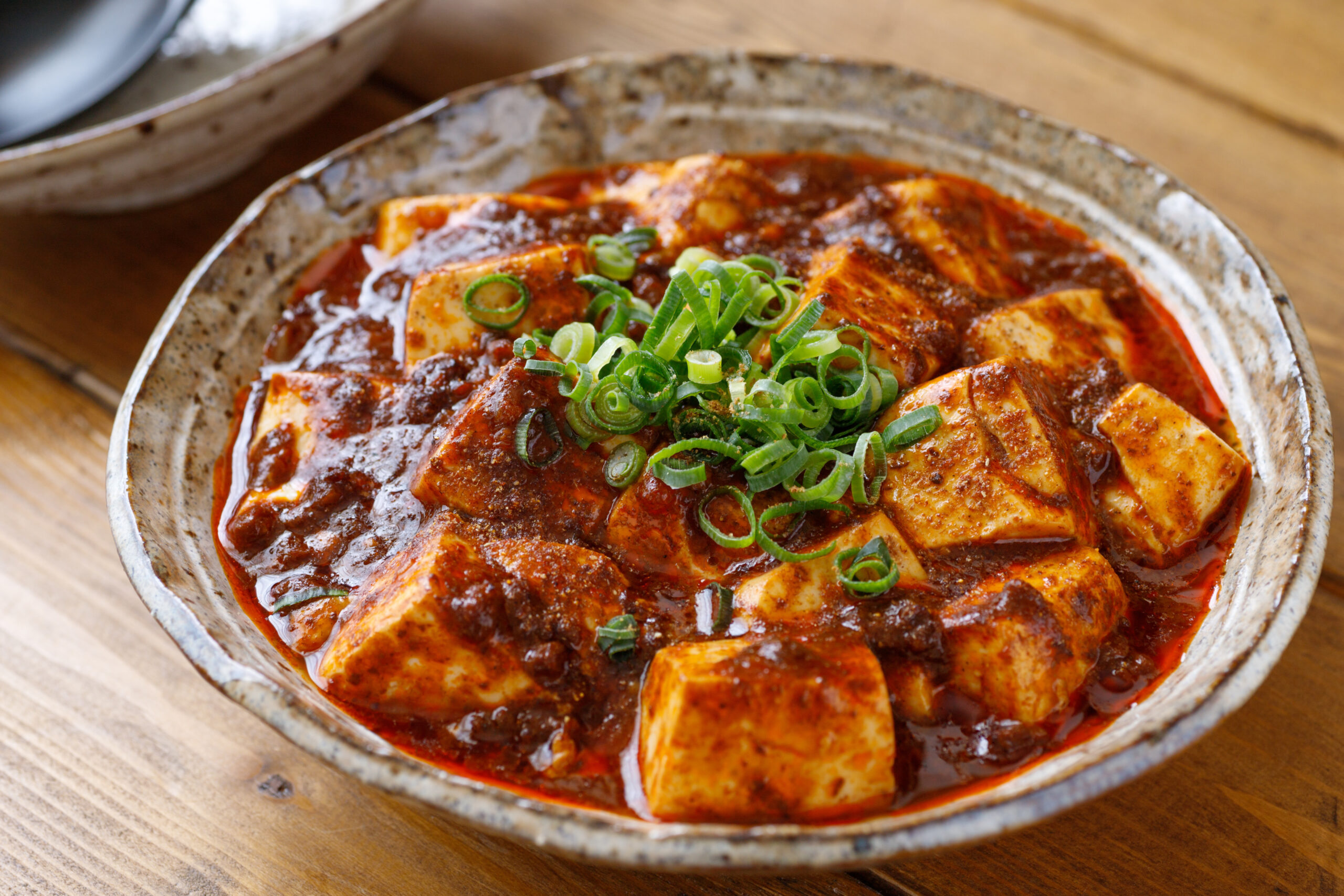 Mapo tofu in a spicy red sauce topped with chopped green onions in a rustic ceramic bowl