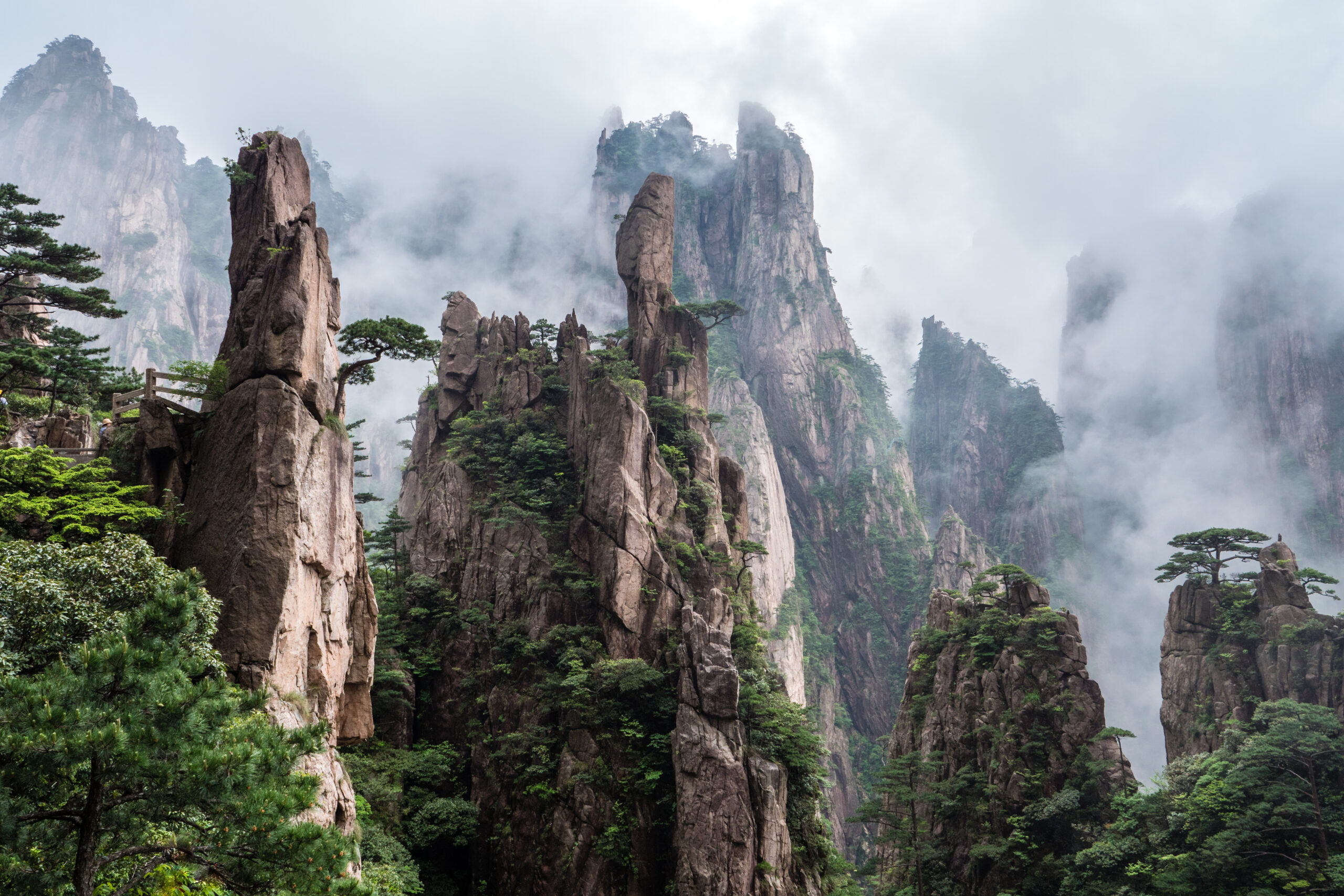Towering jagged rock pillars rise from a misty forest, with green pine trees in the foreground.
