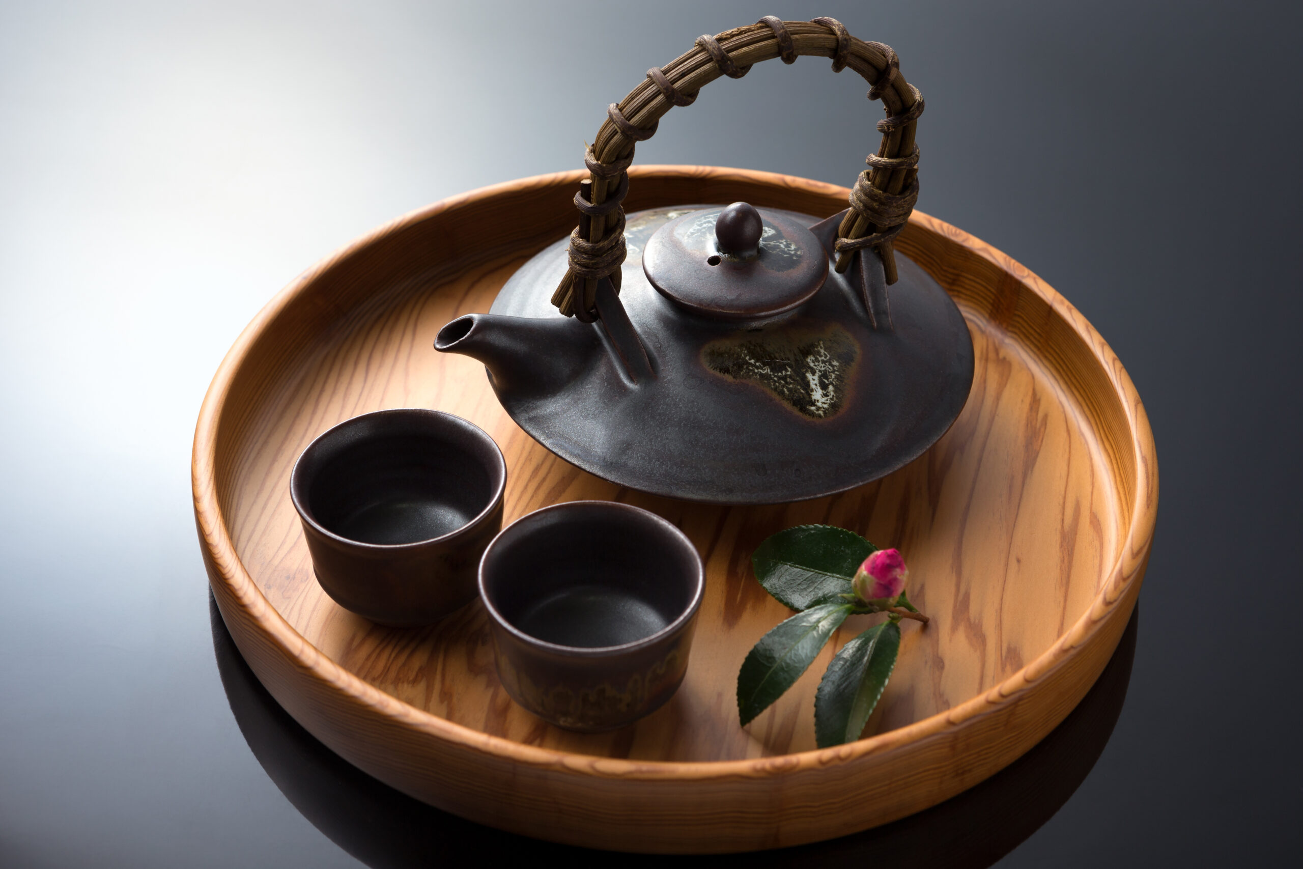 Brown ceramic teapot on a round wooden tray with three small cups and a pink rosebud nearby.