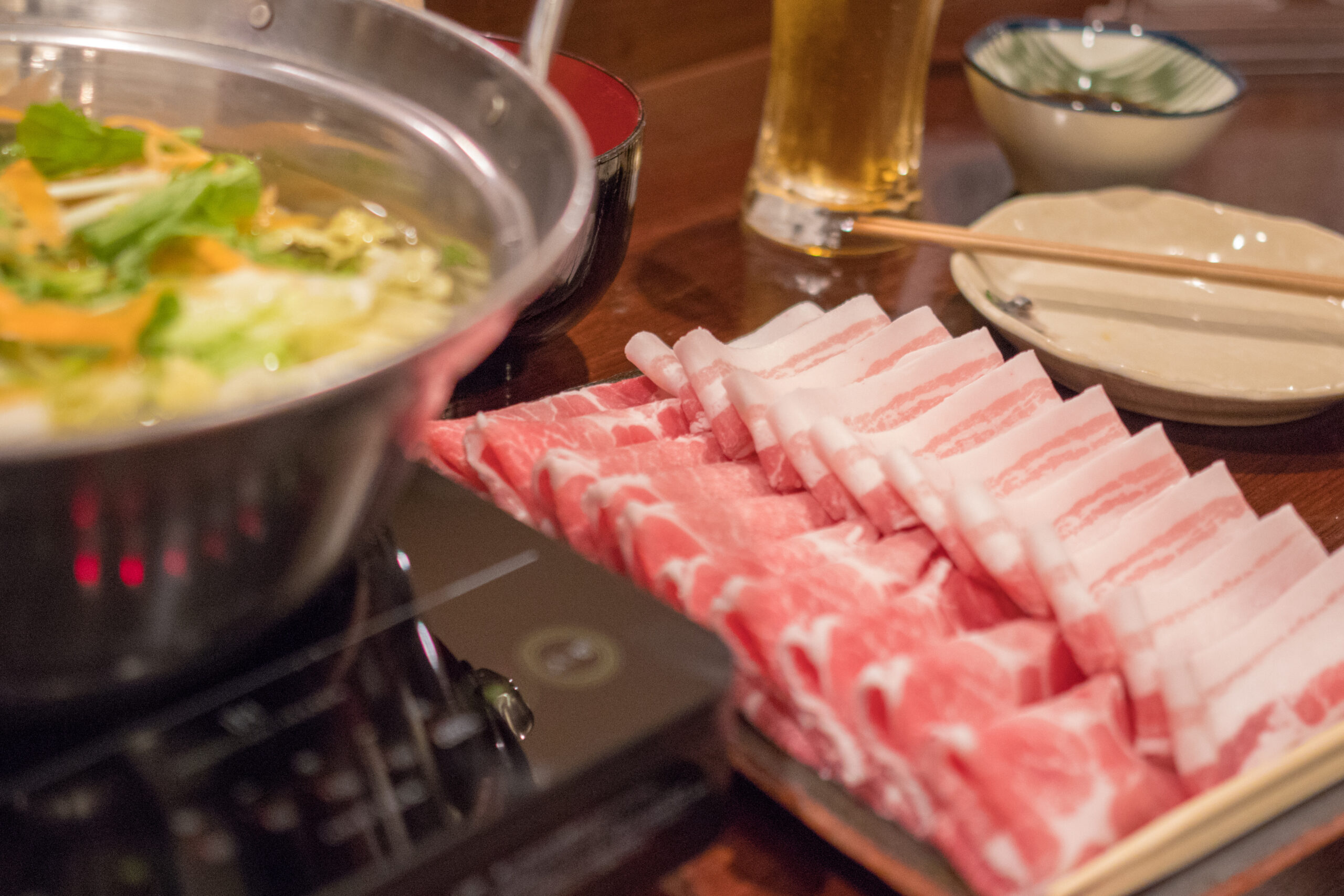Pot of simmering hot pot with greens and vegetables, plus a stack of thinly sliced pork, chopsticks, and a beer on the table.