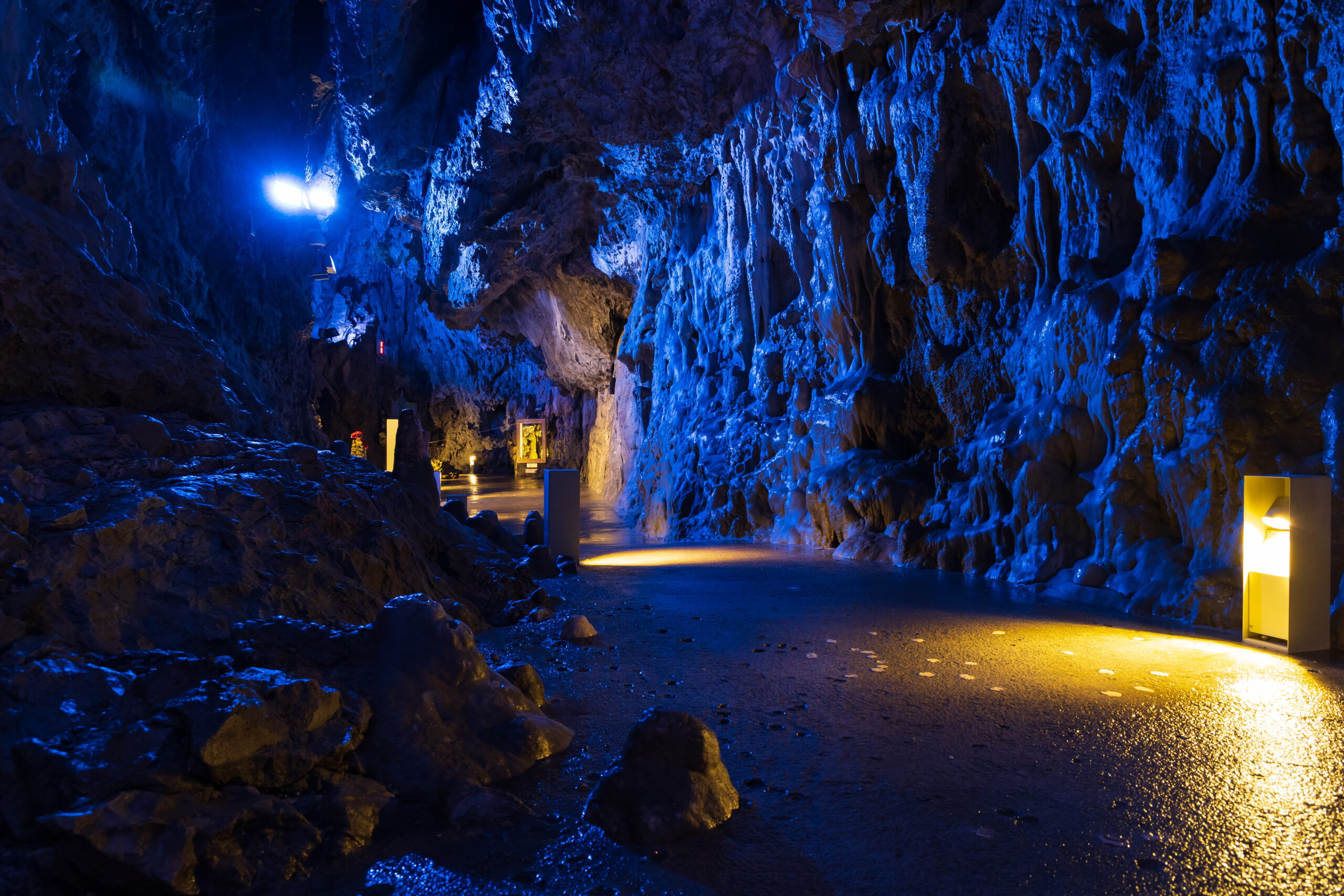 Blue-lit cavern interior with rough rock walls and a paved walkway along the right, illuminated by yellow and blue lights.
